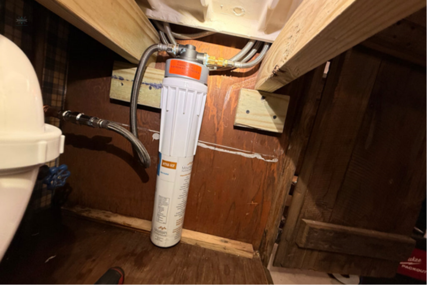 An under-sink water filtration cartridge system installed inside a wooden kitchen or bathroom cabinet.