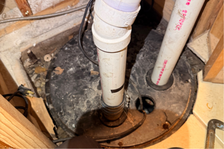 A black sump pump basin cover with white PVC discharge and vent pipes installed in a basement floor.
