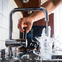 Close-up of a modern chrome kitchen faucet being turned on by a hand; water flows with pressure into a glass, representing Valerio Plumbing's precision plumbing installation and repair services.