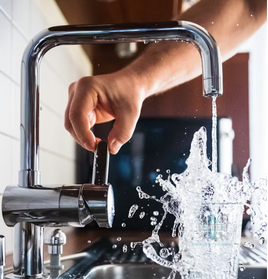 Close-up of a modern chrome kitchen faucet being turned on by a hand; water flows with pressure into a glass, representing Valerio Plumbing's precision plumbing installation and repair services.