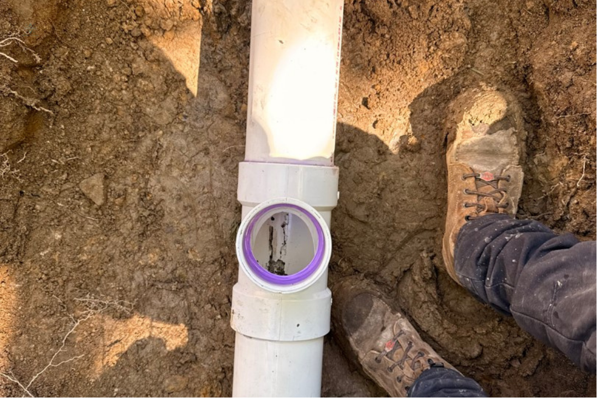 An outdoor white PVC pipe with a T-joint installed underground in a dirt hole next to a shovel.