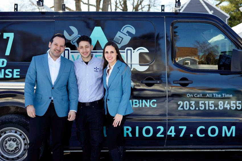 Alejandro, Abel, and Jennifer posing in front of a black Valerio Plumbing service van; the co-owners are wearing blue blazers and Abel is in the gray company shirt, highlighting the logo and 24/7 contact information on the vehicle.