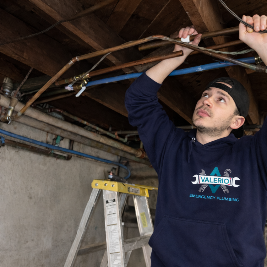 Valerio Plumbing technician Abel Valerio inspecting a network of copper pipes on a basement ceiling; he is wearing a navy blue company hoodie and a black cap, working with precision in a professional residential setting.