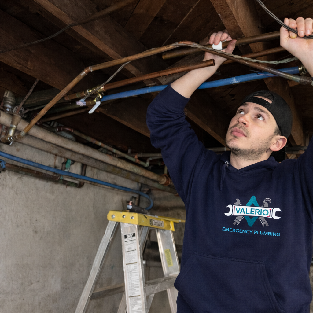 Valerio Plumbing technician Abel Valerio inspecting a network of copper pipes on a basement ceiling; he is wearing a navy blue company hoodie and a black cap, working with precision in a professional residential setting.